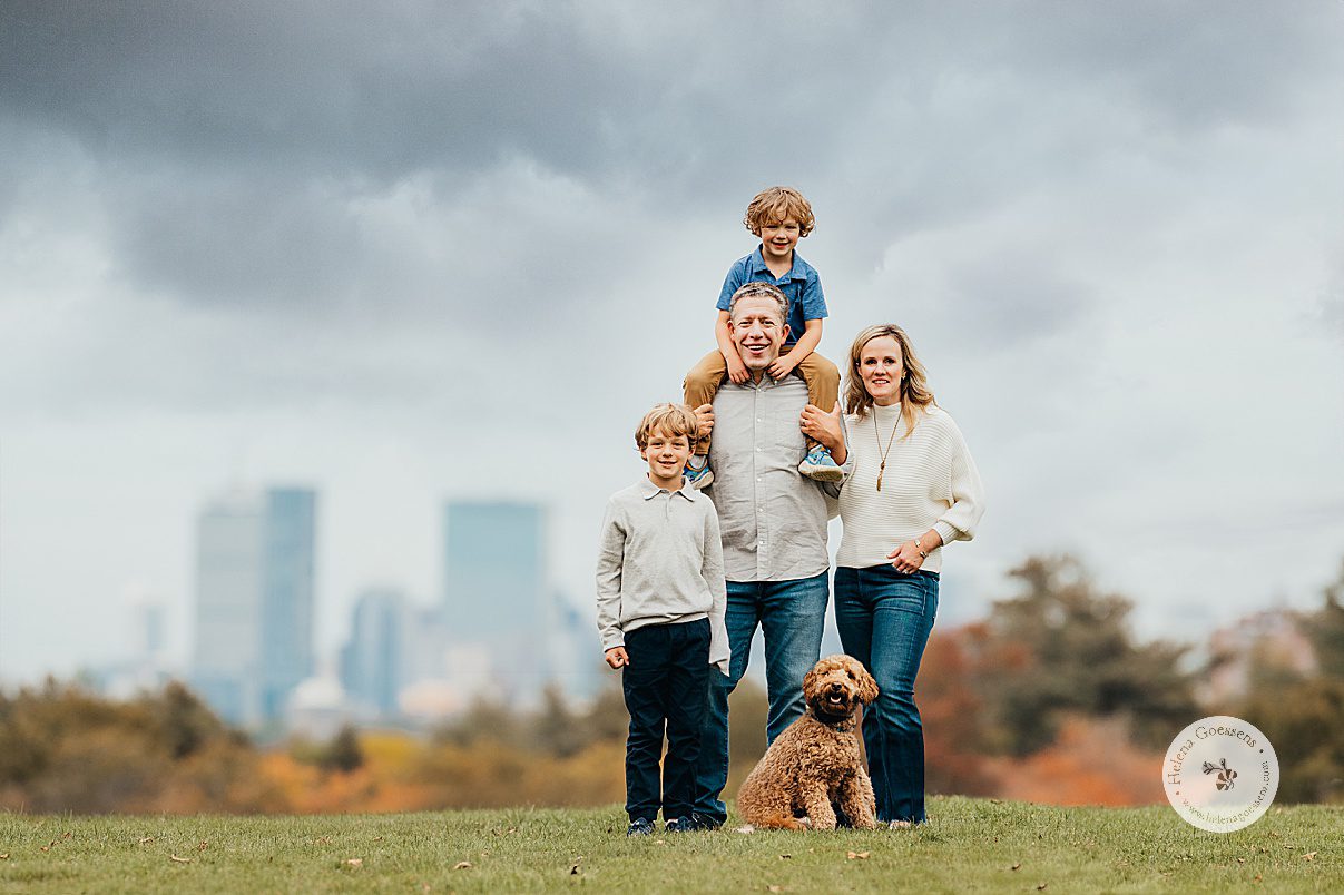 family of four stands with dog in front of Boston skyline in Larz Anderson Park