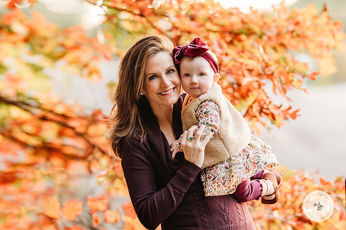 mom in purple sweater holds baby girl in fall dress in front of orange leaves 