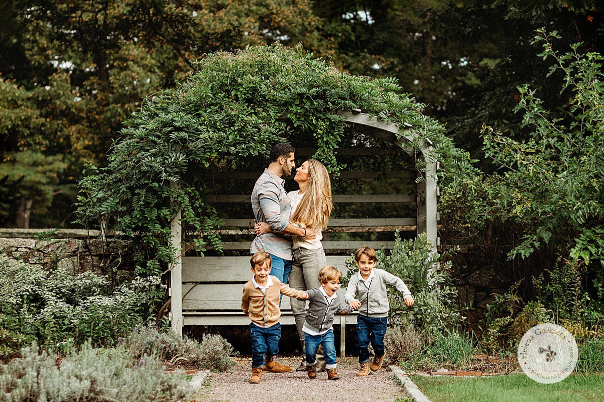 mom and dad lean to kiss while three sons run in front of them during fall family portraits at the Bradley Estate