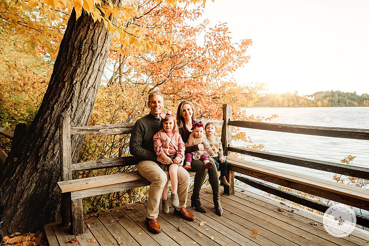 family of five sits on wooden bench near lake during fall family portraits at Wellesley College
