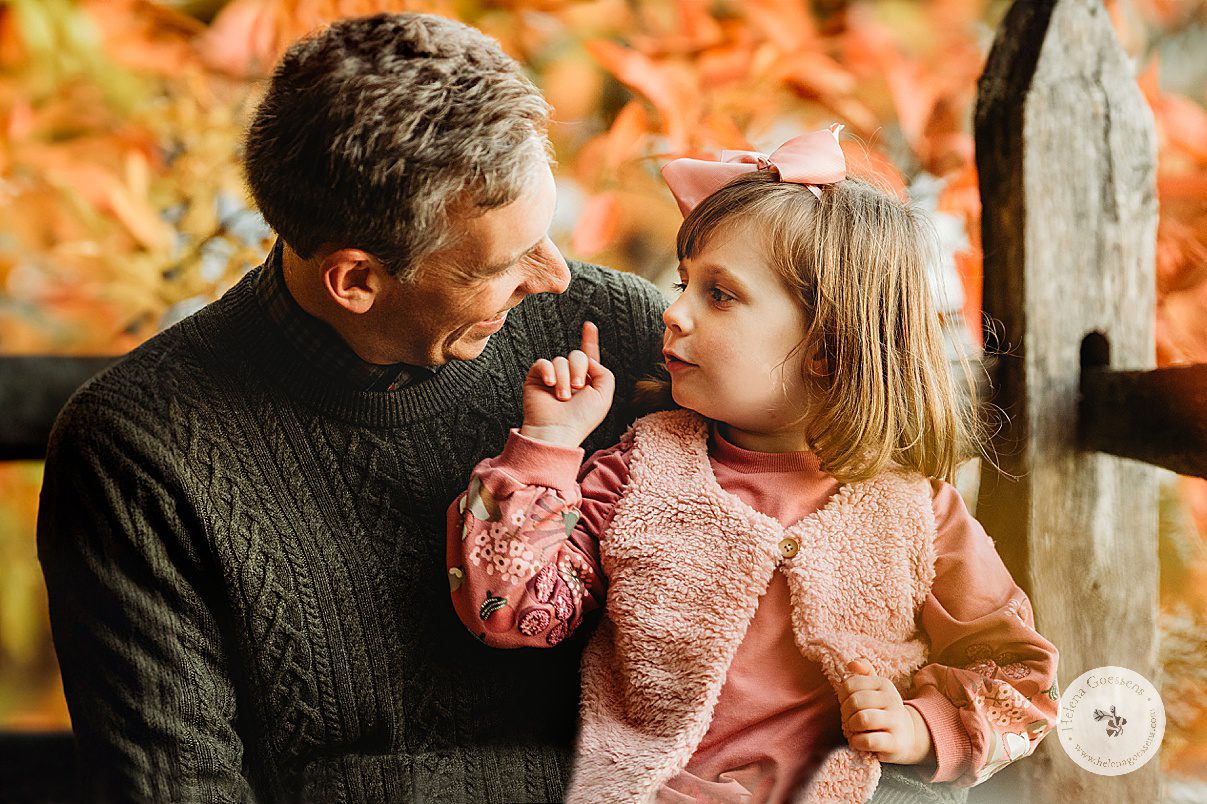 dad sits with daughter in pink dress on his lap by wooden fence during fall family portraits at Wellesley College