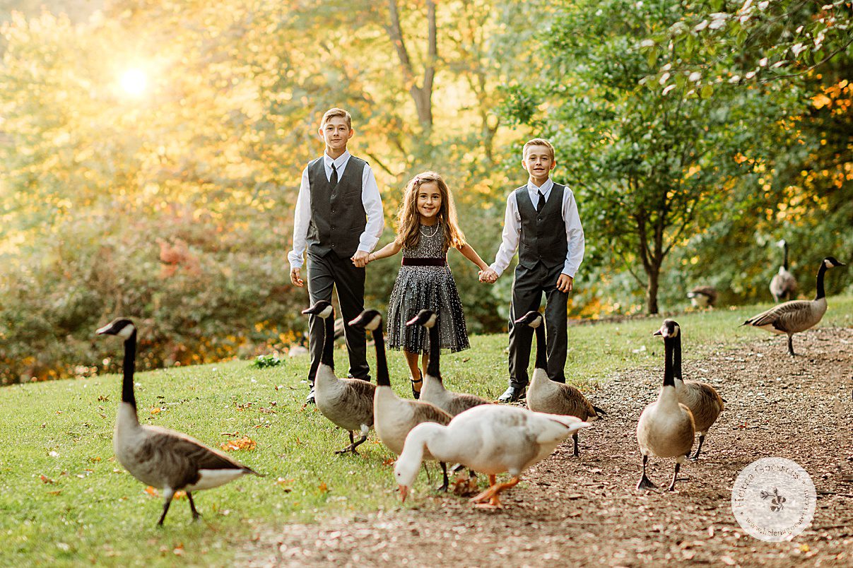 three siblings hold hands walking on path at Wellesley College behind flock of Canadian geese 