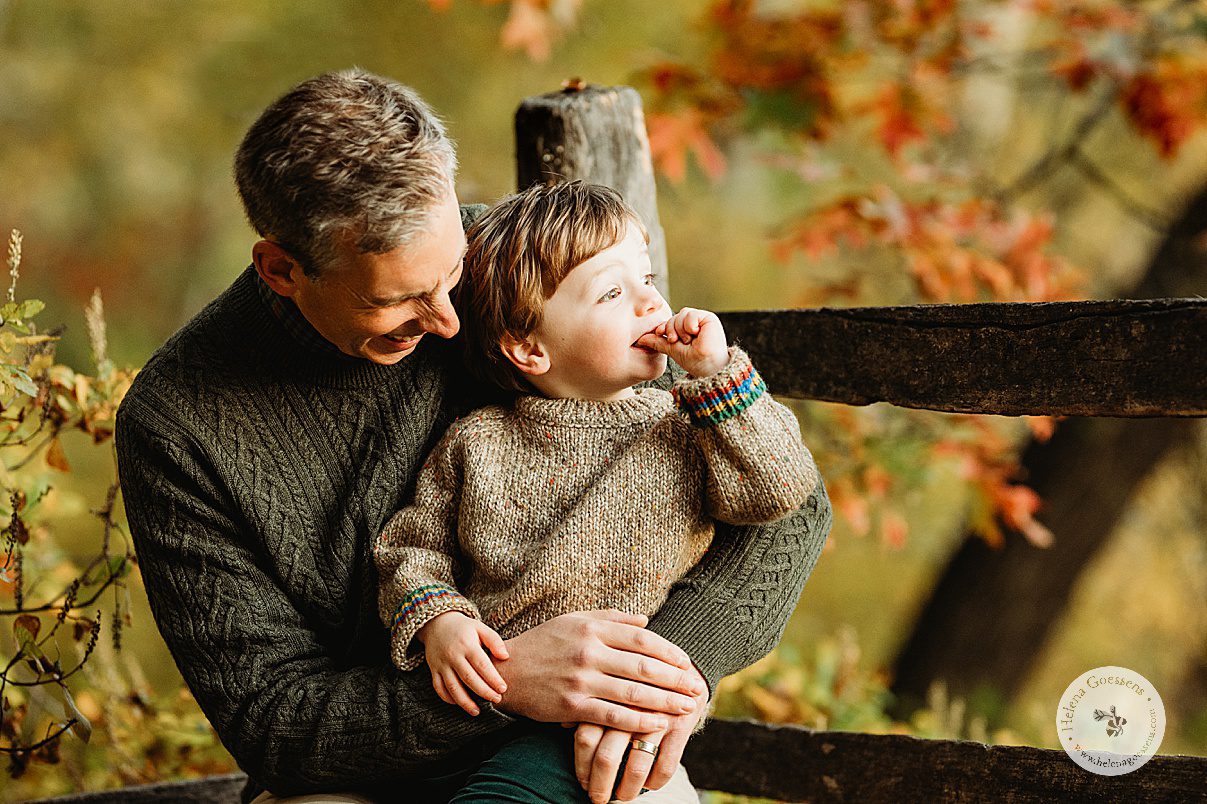 dad in olive sweater holds son in tan sweater along wooden fence during Massachusetts family photos 