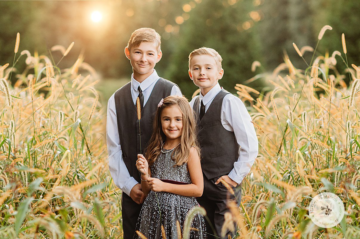 two brothers stand behind younger sister in field of tall grass during family portraits at Wellesley College