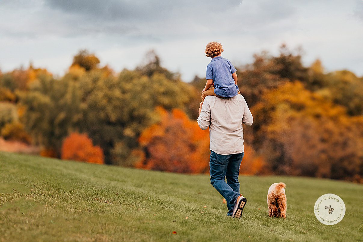 dad walks through Larz Anderson Park with son on shoulders and dog following them 