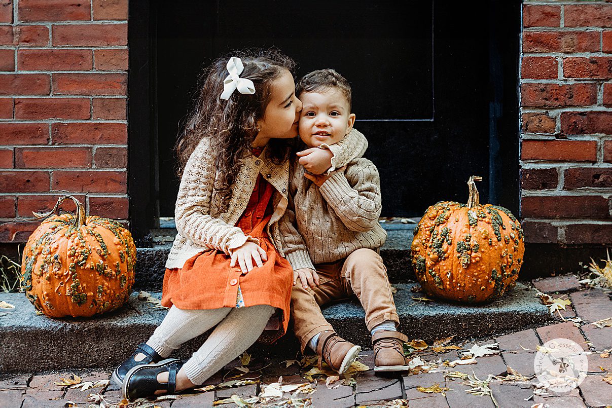 girl in orange dress with tan sweater holds brother's hand and leans to kiss his cheek in front of black door