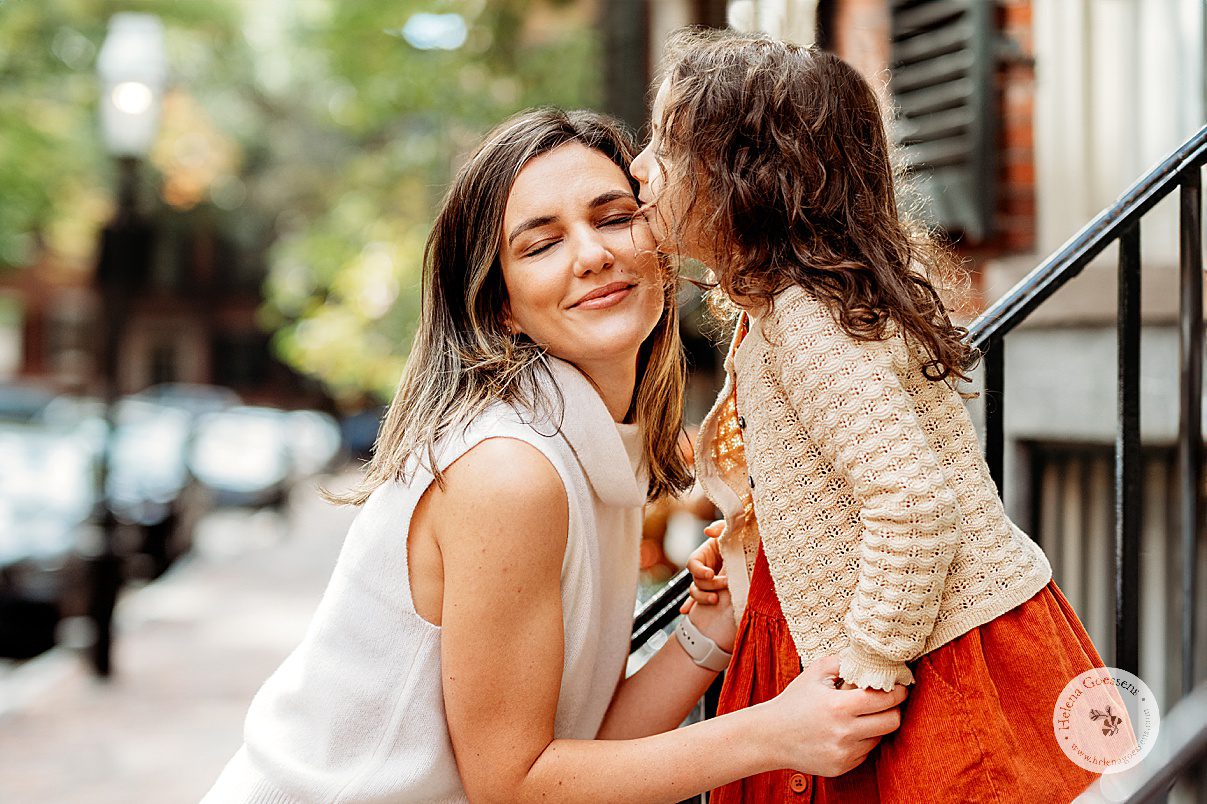 girl in orange dress leans to kiss mom's forehead during family photos on Acorn Street on Beacon Hill in Boston MA 