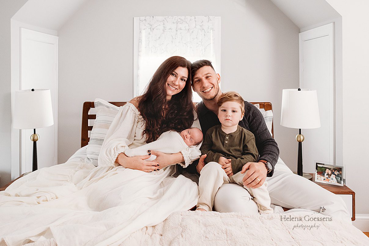 parents sit on bed with two young sons during lifestyle newborn portraits in Waltham MA