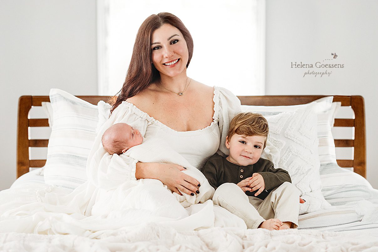 mother in white dress holds baby boy and toddler son on bed during lifestyle newborn portraits in Waltham MA