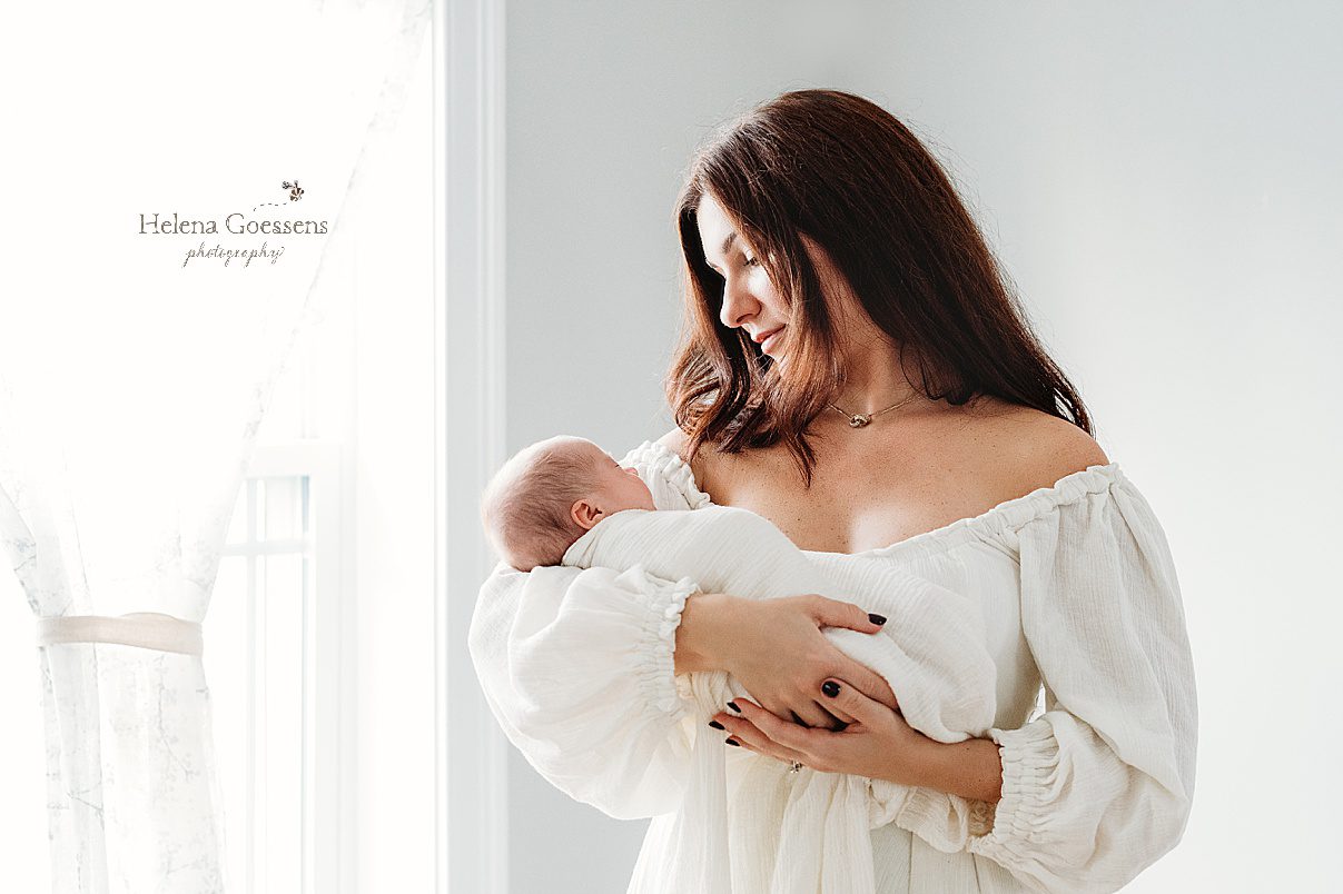 mother in white off-the-shoulder gown holds baby boy in arms during during lifestyle newborn portraits in Waltham MA
