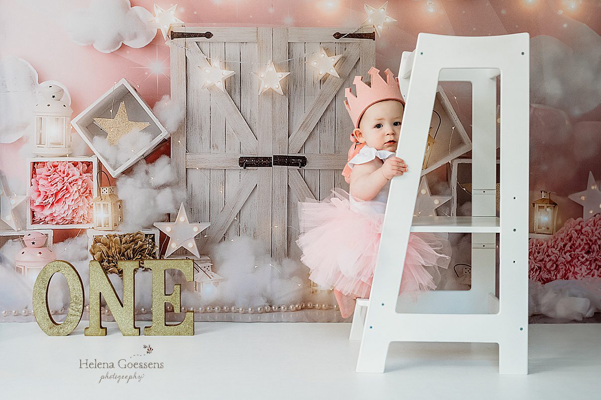 one year old in pink crown and tutu climbs on step stool during first birthday Twinkle Twinkle Little Star cake smash with pink, gold, and white details 