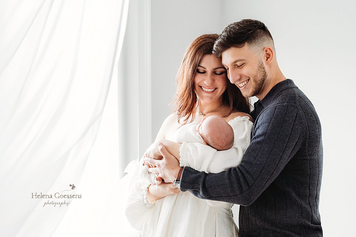 parents smile down at newborn son by window and white curtains during lifestyle newborn portraits in Waltham MA