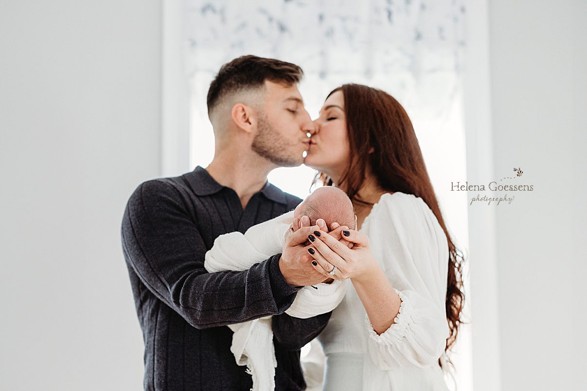 parents kiss while holding son out in front of them during newborn photos at home 
