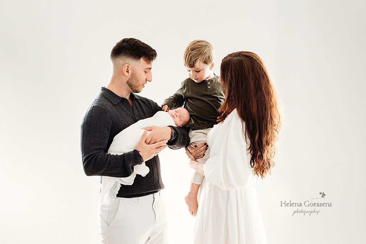parents pose holding two sons in front of white wall while big brother touches baby's cheek