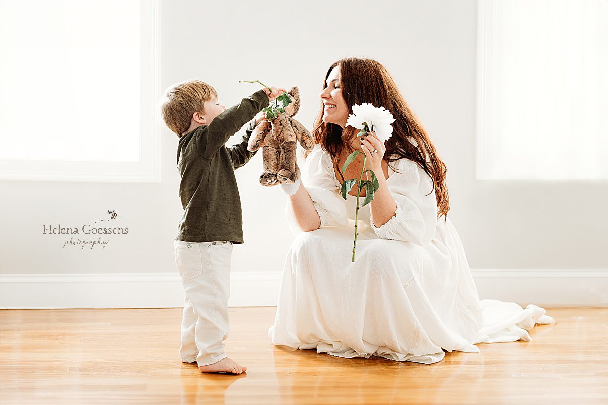 mom kneels down to play with flowers and bear with son during lifestyle newborn portraits in Waltham MA