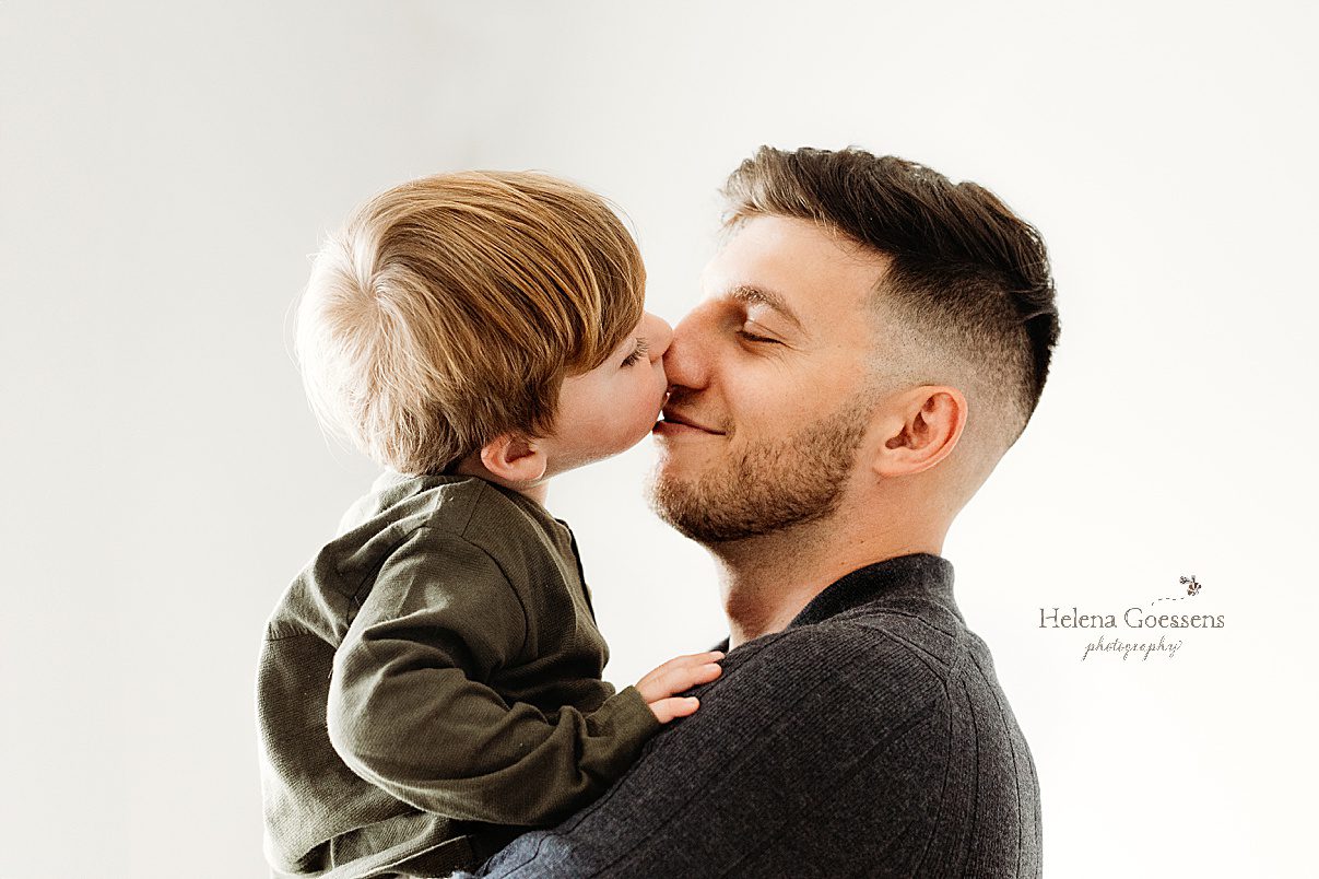 dad holds toddler son nuzzling noses in front of white wall