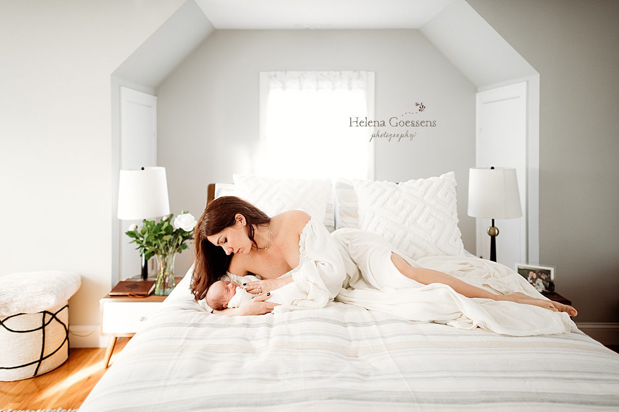 mom lays in boho white dress with son in crook of arm on bed during lifestyle newborn portraits in Waltham MA