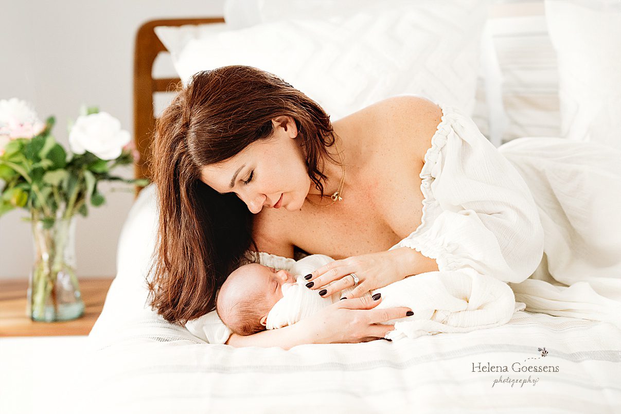 mother in off-the-shoulder white gown looks down at baby boy in arms during lifestyle newborn portraits in Waltham MA