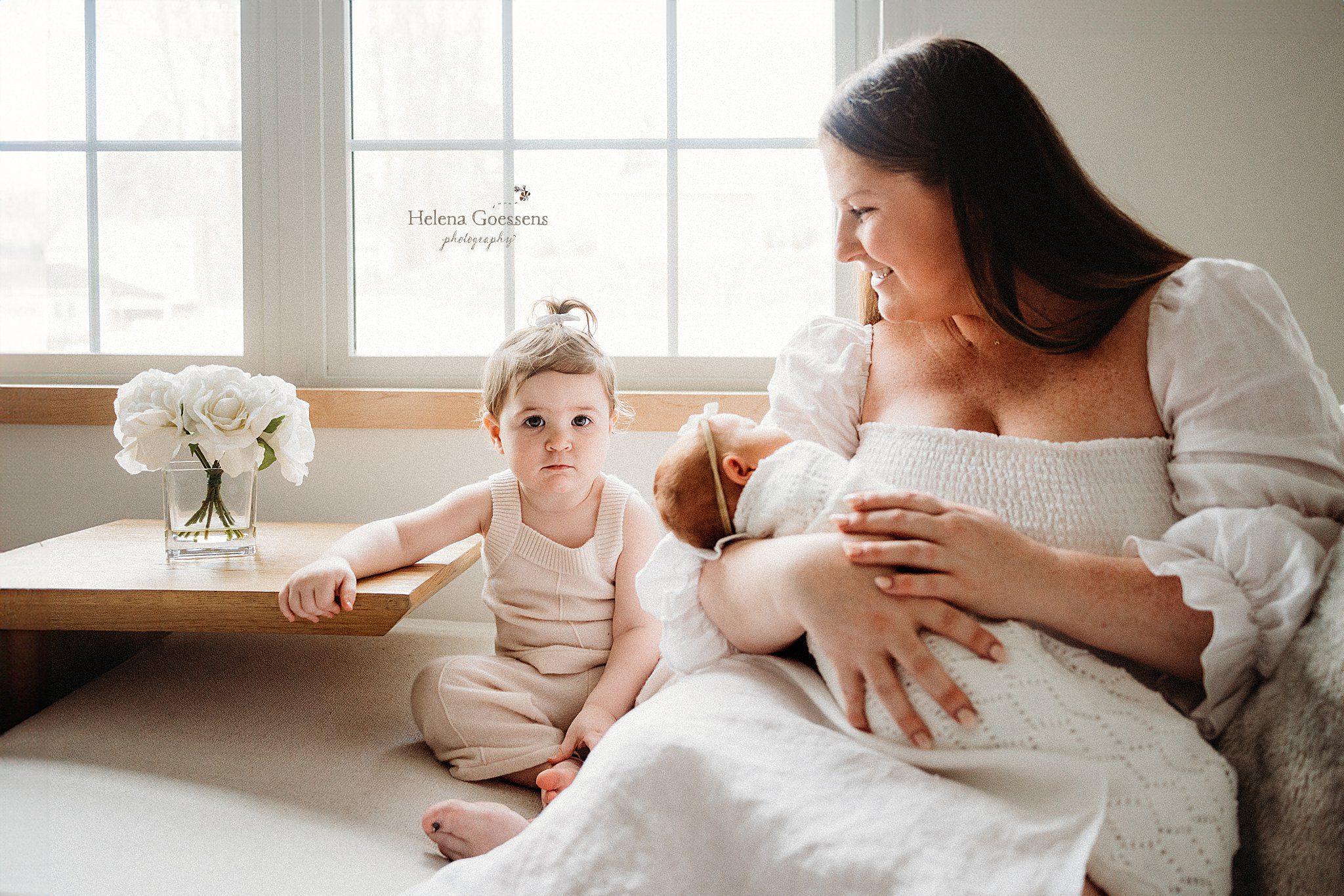 mother in white dress smiles at toddler daughter and holds newborn during in home newborn portraits in Canton MA