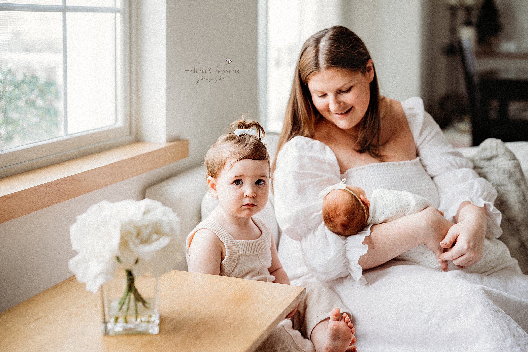 mom holds newborn daughter and looks at toddler next to her during at home newborn portraits in Canton MA