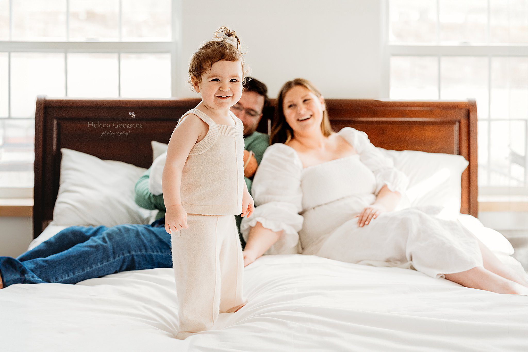 toddler stands on parents' bed smiling at camera during lifestyle family photos 