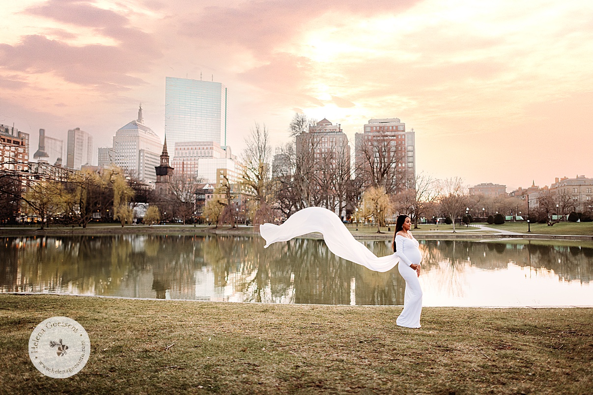 expecting mother in white dress holds baby bump standing in front of pond in Boston Public Garden at sunset 