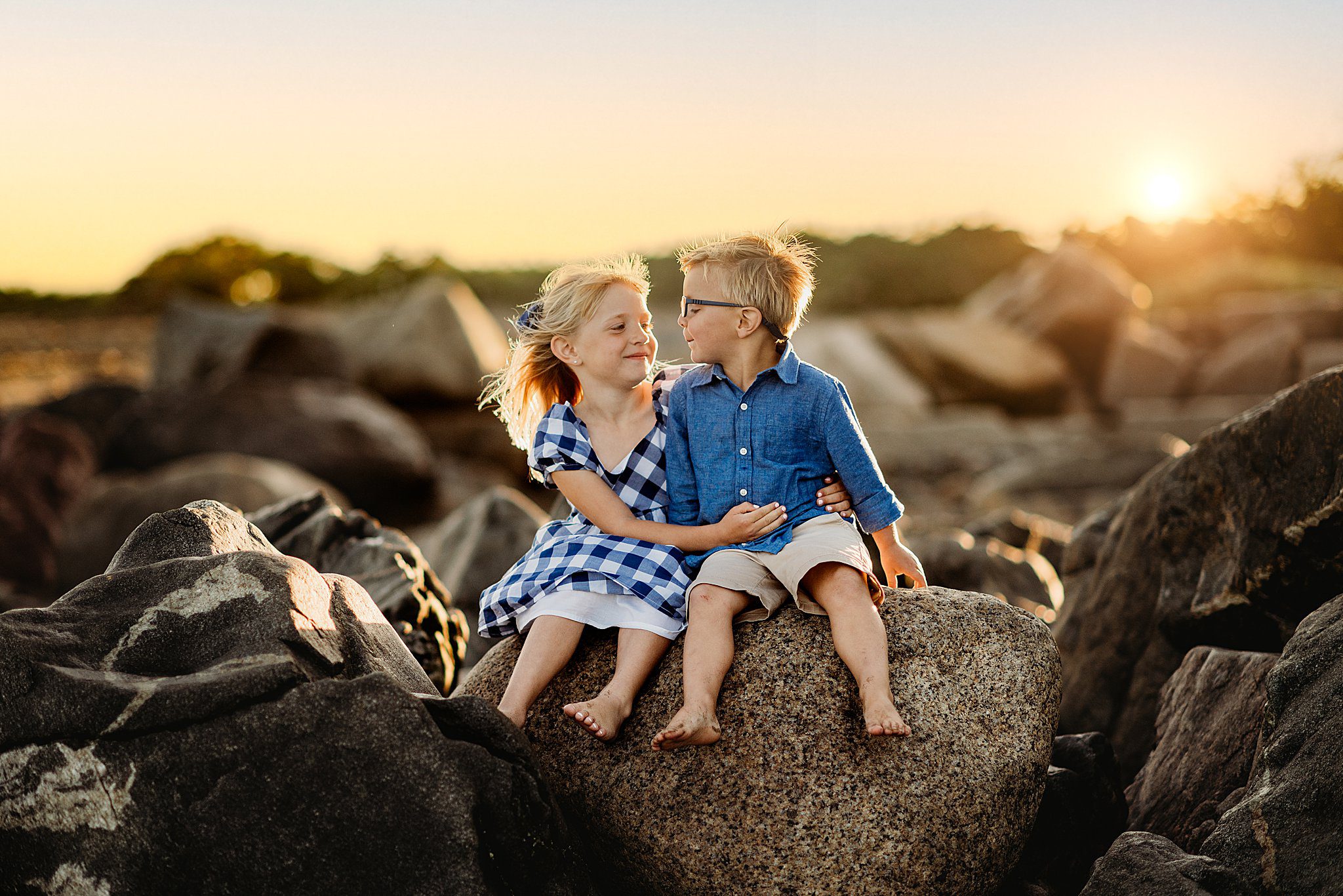 Two children sit on a large rock at sunset. The girl in a blue checkered dress smiles at the boy in a denim shirt and shorts. A Boston photographer captures this idyllic moment, where surrounding rocks and the setting sun create a warm backdrop perfect for family photography.