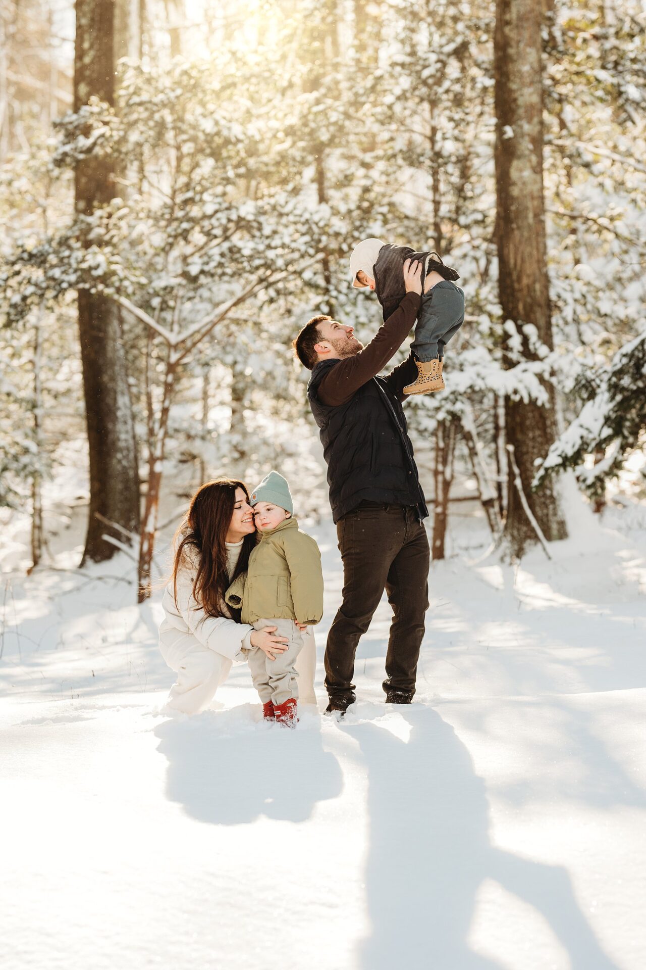 A Boston Family Photographer captures a joyful family in a snowy forest. A woman crouches, hugging a toddler, as a man lifts another child into the sunlit air—creating a warm, timeless moment.