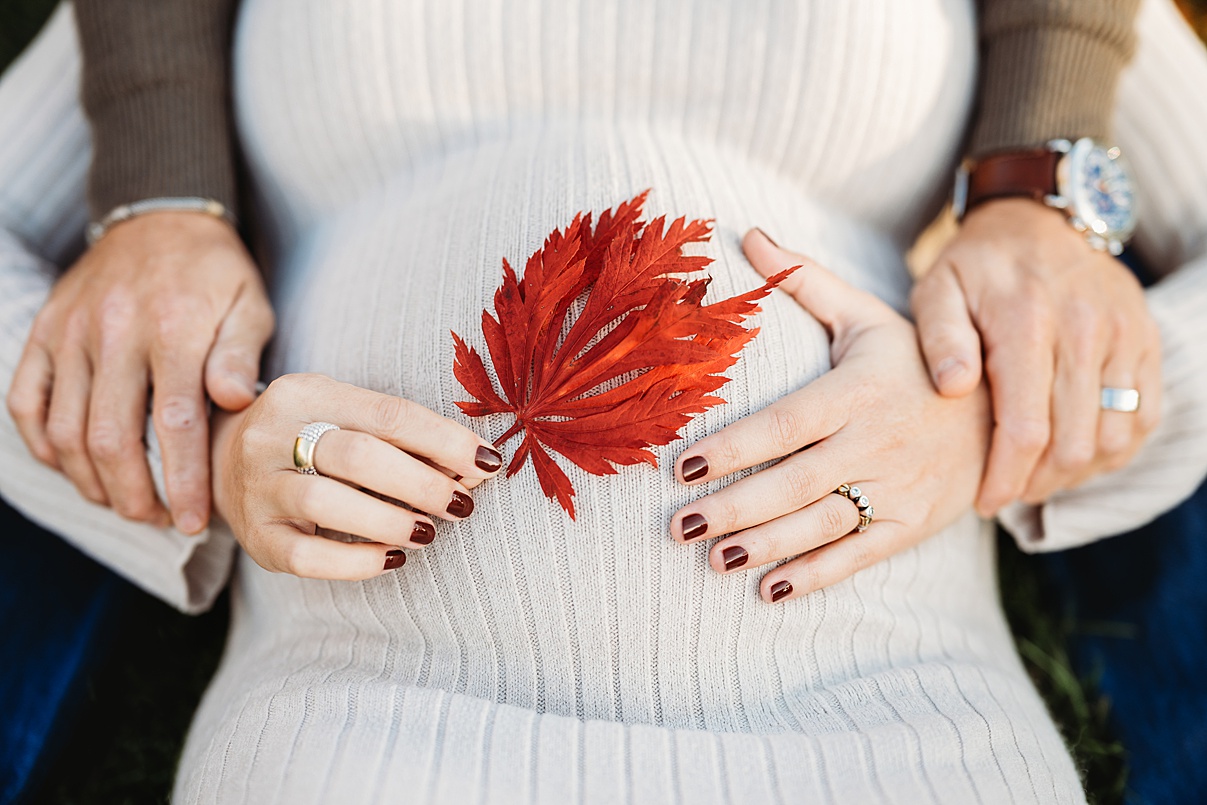 A pregnant person wearing a cream sweater holds a red autumn leaf on their belly, with another’s hands gently resting on their sides. Both have rings on their fingers—capturing the warmth of family moments with a Lifestyle Boston Photographer.