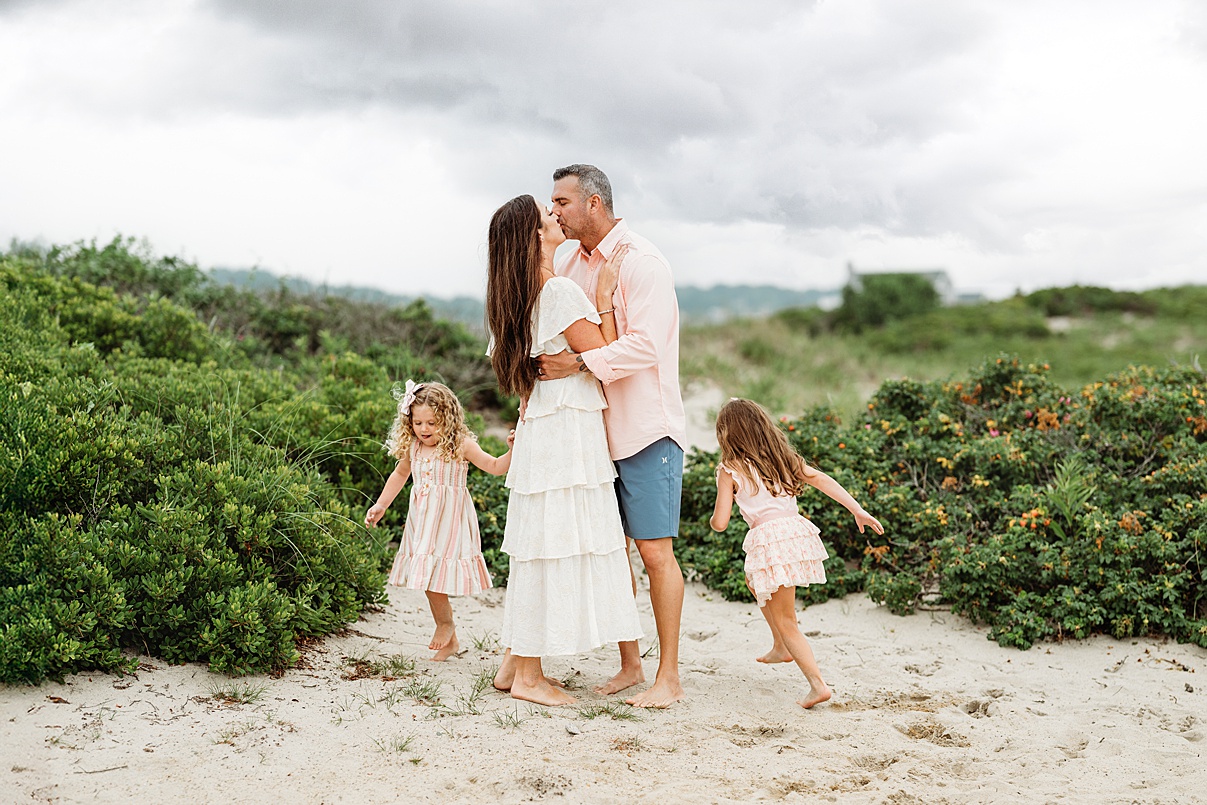 A couple kisses on a sandy beach, surrounded by greenery, while two young girls in light dresses play nearby. The cloudy sky adds softness to this Lifestyle Family Photography moment captured by a Boston Family Photographer.