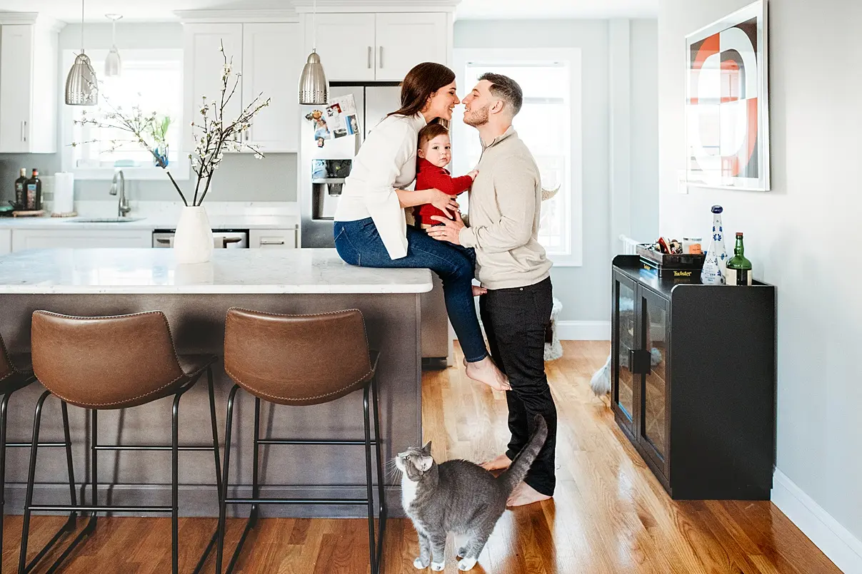 A couple with a child sits on a kitchen counter. The man kisses the woman's forehead while holding the child. A cat stands on the floor. Kitchen features stools and modern decor, captured perfectly by a Boston Newborn and Lifestyle Family Photographer.
