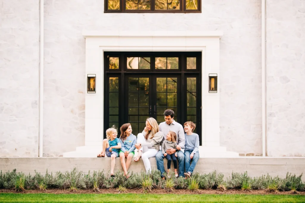 A family of six, including four children, sits together on a ledge in front of a house with large black-framed windows and a stone exterior, perfectly captured by a Boston Newborn and Family Photographer.