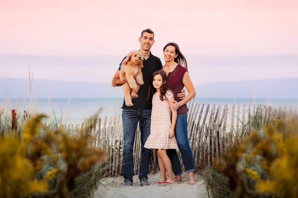 A family of four, including a man, woman, girl, and their dog, stand on a beach path with dune grass in Massachusetts. Smiling at the camera under a pink sky with the ocean behind them, they capture a perfect portrait moment thanks to our professional portrait service.