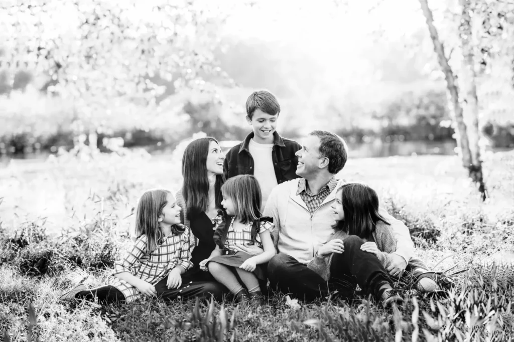 A family of six sits on grass, smiling and looking at each other. Three adults and three children are present. The background is a sunlit, blurred outdoor setting. The image is in black and white.
