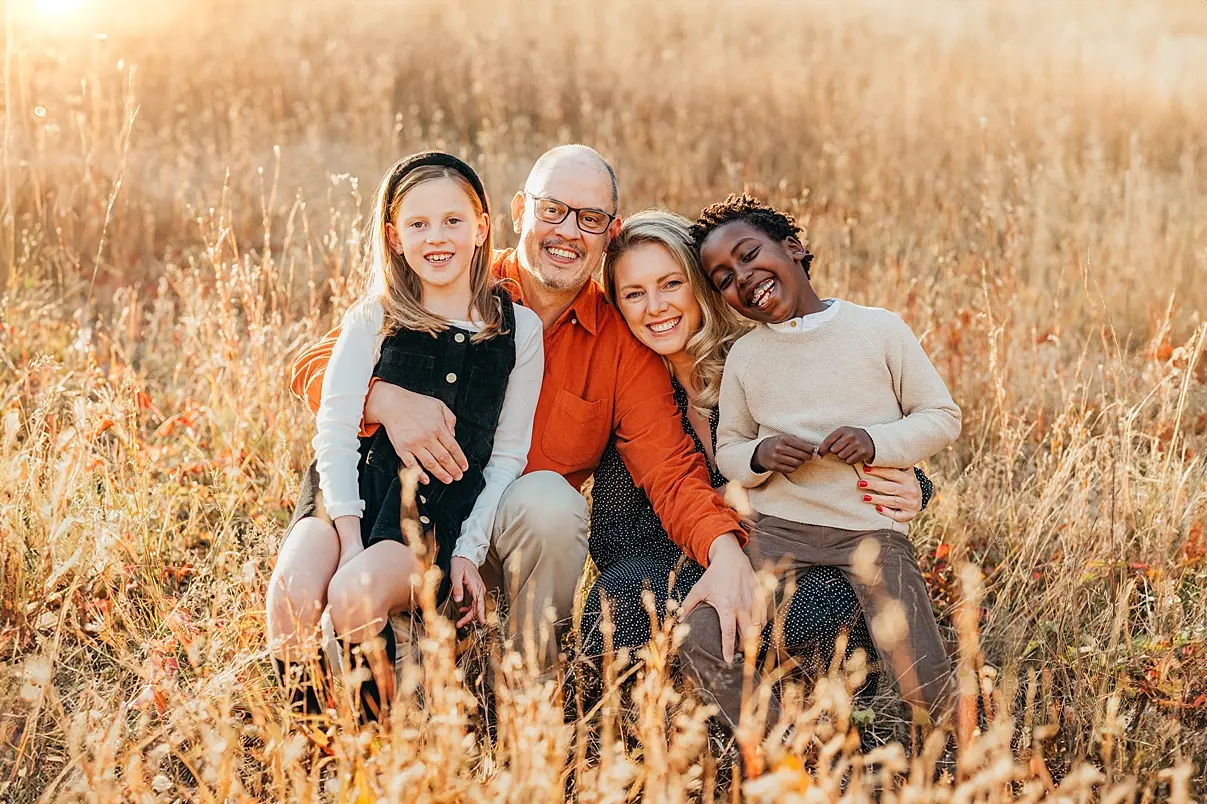 Family of four sitting and smiling in a sunlit field, with tall grass surrounding them.