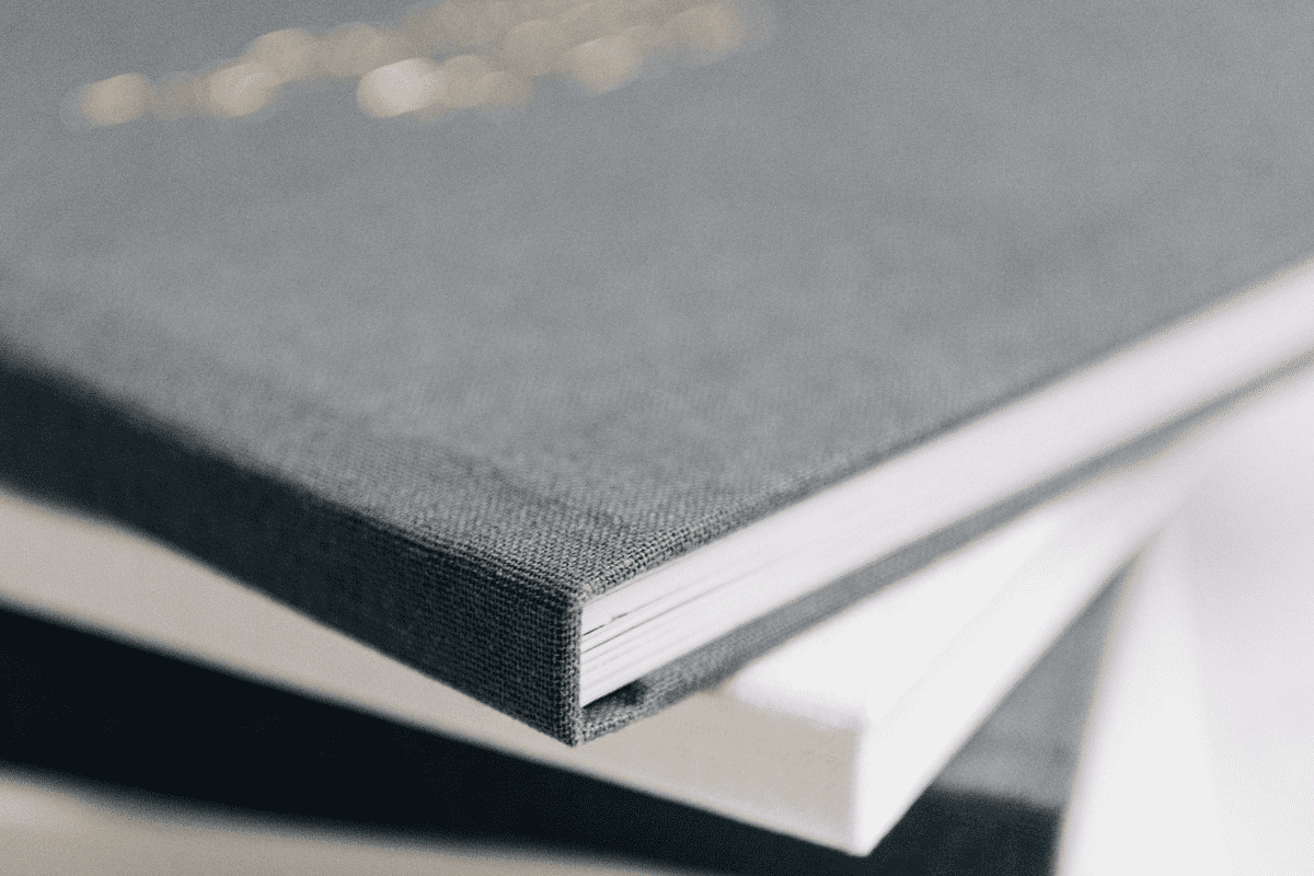 Three hardcover books with textured covers are stacked at an angle. The top cover is dark gray, the middle one is dark blue, and the bottom one is white.