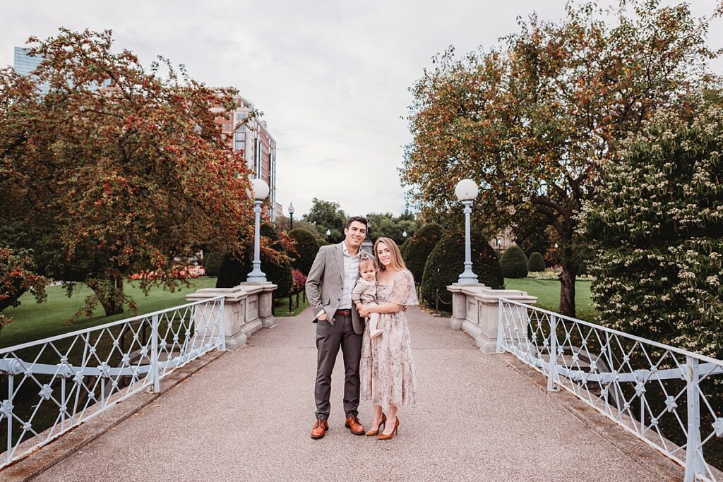 The couple stands on a decorative bridge in the park, cradling their baby. Surrounded by trees with vibrant leaves, they embrace the serenity of an overcast day. The man, in a gray suit, and the woman, in a floral dress, capture this perfect Spring Family Session moment.