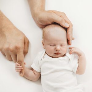A newborn baby sleeps peacefully on a white surface, holding an adults finger. The adults hand gently rests on the babys head. The baby is wearing a white onesie, creating a serene and tender scene.