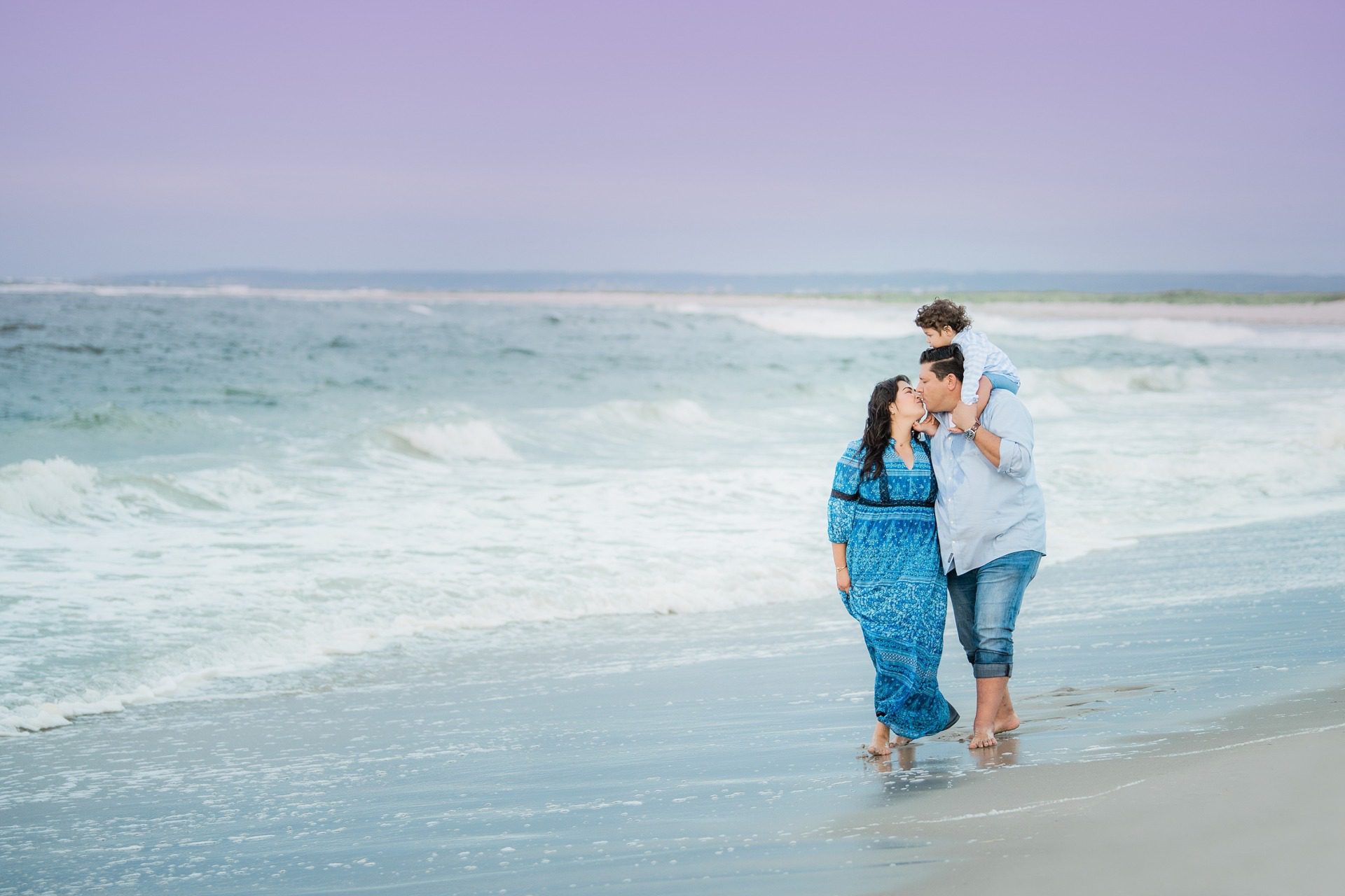A couple walks hand in hand along a beach, with the man carrying a child on his shoulders. They are barefoot, enjoying the waves and the scenic, calm seaside under a soft, purple tinted sky.