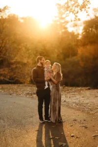 A couple stands on a path during sunset, holding a small child. The sun is setting behind them, casting warm light and long shadows. Autumn foliage is visible in the background.