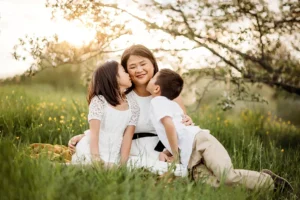 A woman sitting on grass with two children, one on each side, in a field. The children are leaning in to kiss her cheeks. It's a sunny day with trees and flowers in the background.