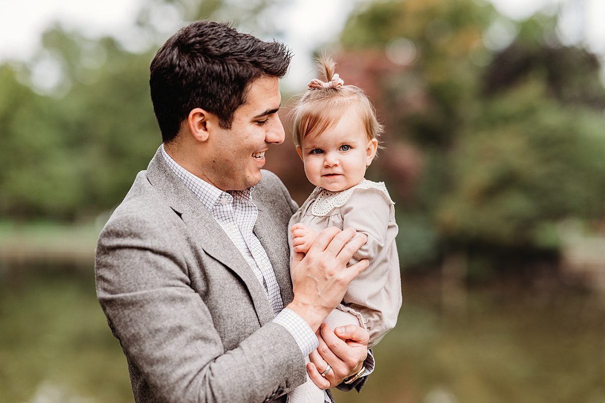 A man in a gray suit holds a young child with a small ponytail, dressed in a beige outfit. They are outdoors, with blurry green trees and water in the background, both smiling and looking content.