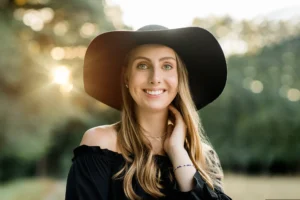 Woman smiling outdoors, wearing a wide brimmed black hat and black top, with sunlit trees in the background.