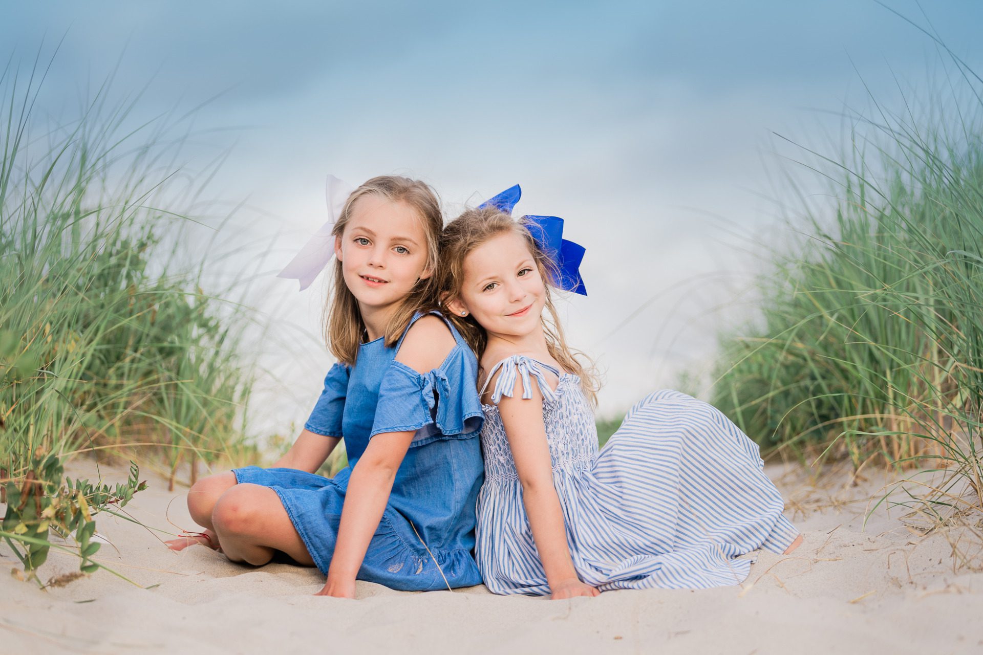 Two young girls sitting back to back on a sandy beach path surrounded by grass. One wears a blue dress with a white bow, and the other wears a striped dress with a blue bow. They smile with a cloudy sky in the background.