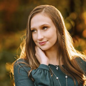 A woman with long brown hair is outdoors, wearing a green top. She is touching her neck and looking to the side, with a gentle smile. The background is blurred with warm, golden autumn colors. A silver necklace is visible around her neck.