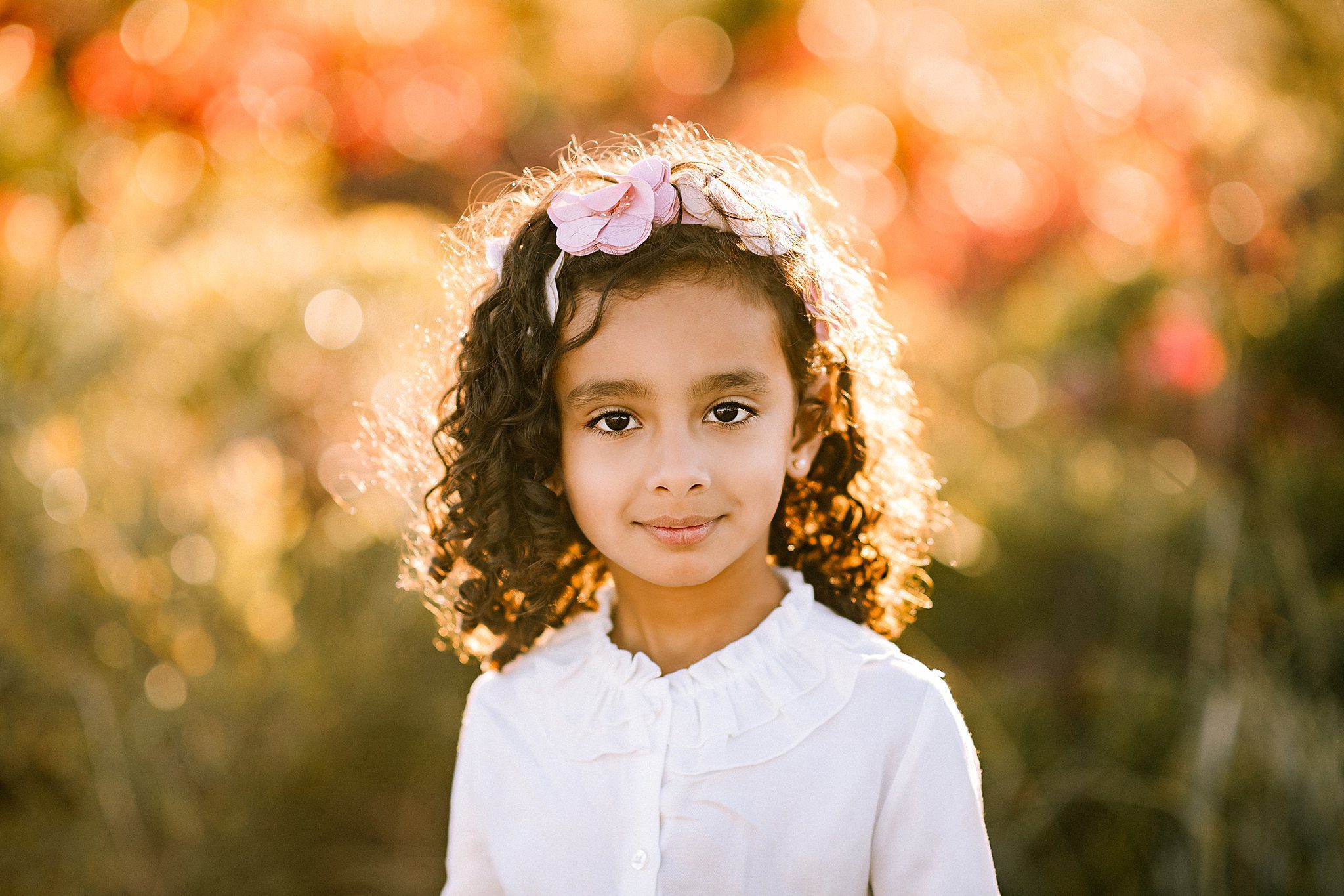 A young girl with curly hair and a pink flower headband smiles softly. She wears a white ruffled blouse. The background is a warm, blurred outdoor setting with sunlight filtering through, creating a bokeh effect.