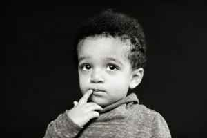 A child with curly hair looks upward thoughtfully, touching their lips with a finger. The background is dark, and the child is wearing a light colored sweater.