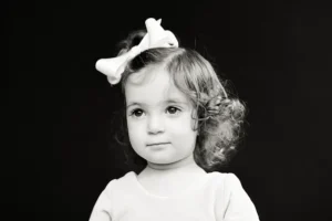 Black and white photo of a young child with curly hair and a large bow, looking slightly to the side against a dark background.
