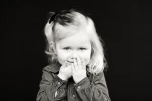 Black and white photo of a young child with light hair, wearing a denim shirt and a hair bow, covering their mouth with both hands against a dark background.
