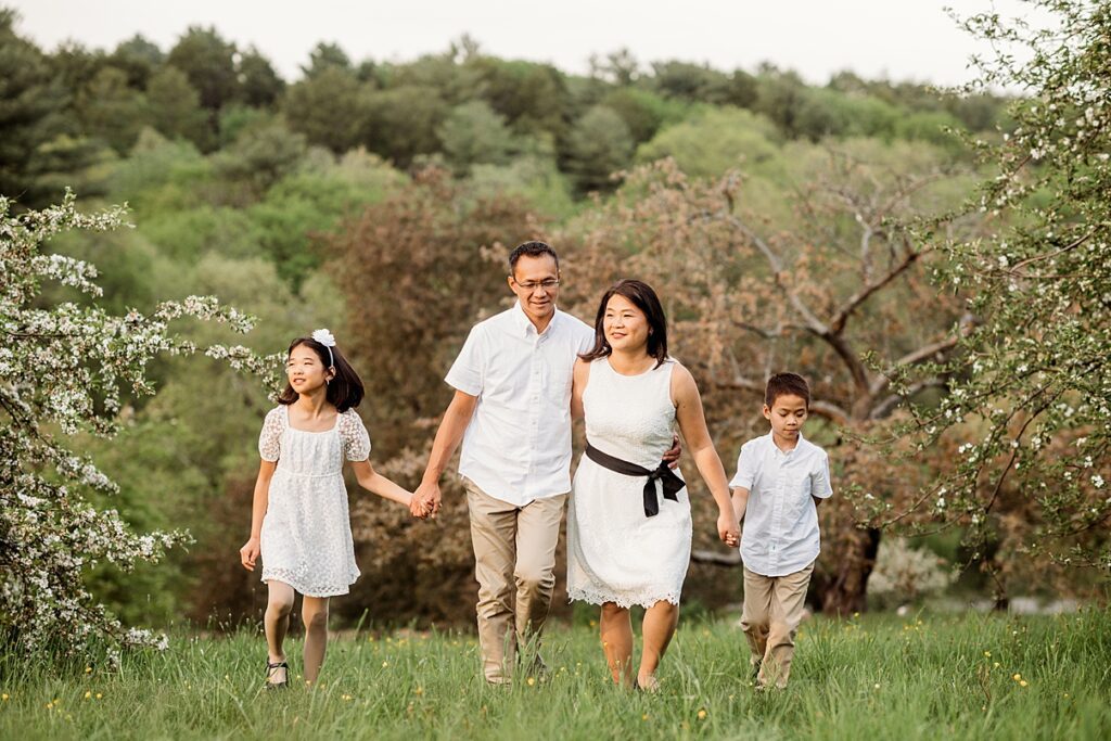 A family of four, dressed in white and beige, walks hand in hand through a grassy field with trees in the background. They appear happy and are enjoying a sunny day during their spring family session.